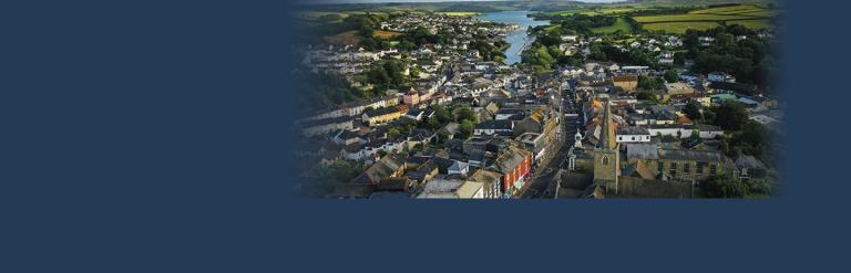 An aerial view of Kingsbridge Fore Street, taken in the early morning. In the distance the estuary is visible and lit by the morning sun. Most of the buildings are still shaded. 