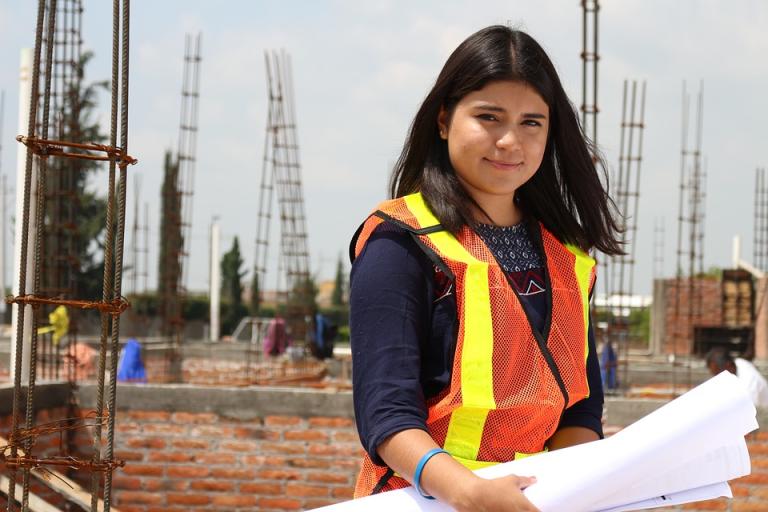 A photograph of a young woman with long dark hair, looking over plans and blueprints. A building site is visible in the background.