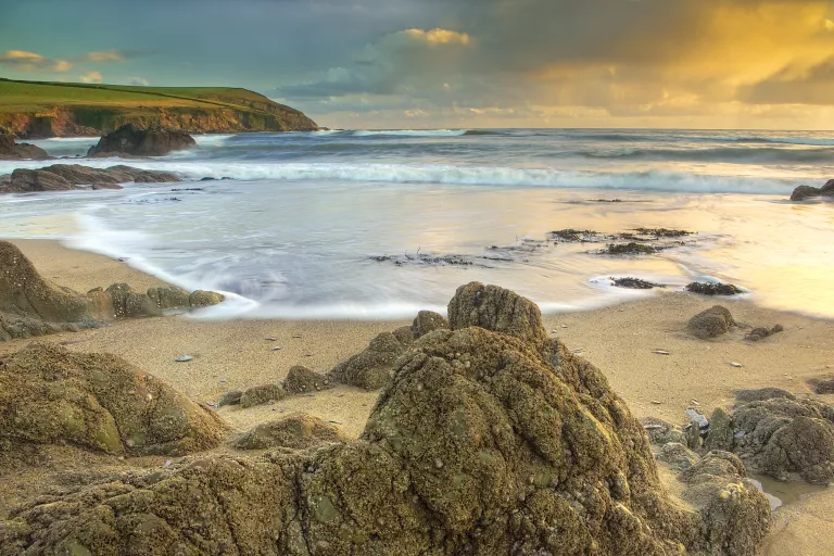 A photograph of the beach at Hope Cove at sunset. In the foreground, jagged rocks and sands are washed by the sea, which is quite choppy with white foam on the waves. The sky is blue but there are some fluffy clouds which reflect the orange light from the setting sun.