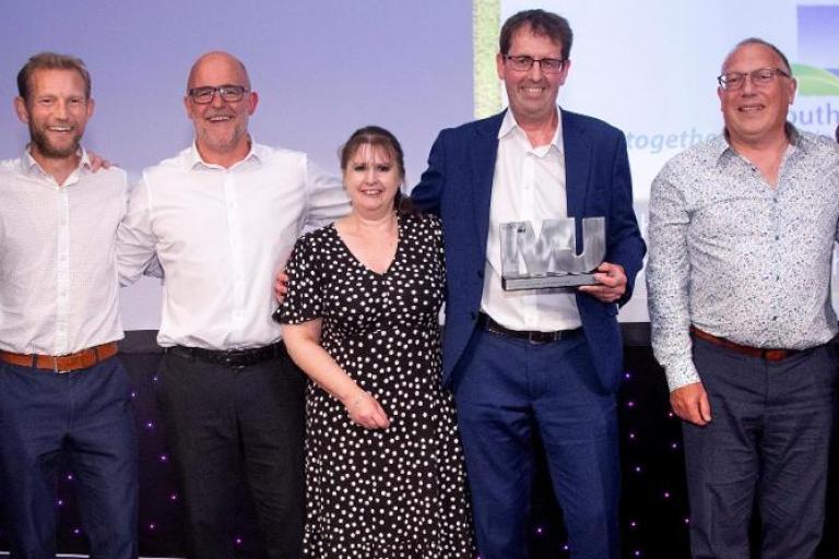A photograph of the five members of the senior leadership team receiving their award. They are smiling broadly at the camera and have their arms around each other's shoulders.