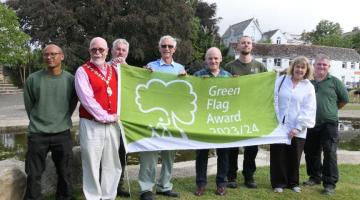 A group of eight people proudly holding up the new green flag awarded to Kingsbridge recreation ground. The park and its pond are visible in the background.