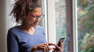 A young black woman, wearing glasses and blue top, is using her phone while standing next to a window. She's smiling slightly, as if she's found an answer she was looking for.