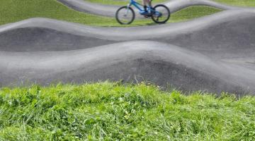 A person on a sports bike rides over a pump track, which is a bike track with lots of little hillocks for jumping and stunts. There is green grass between the different sections of the track.