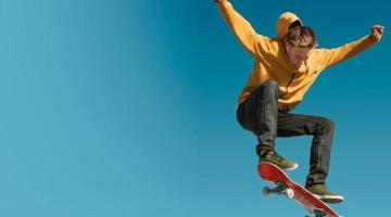A young man in a yellow hoody performs a jump on skateboard. The background is blue, suggesting he's leaping high into the sky.