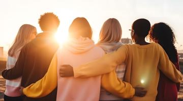 Six young people, with their backs to the camera, are looking at a bright summer sunrise. The sky is blue. Each of the six people is locked together in a gentle uniting embrace, with their arms around each other's backs.
