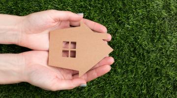 hands holding a brown miniature house with grass background