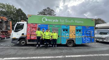 Four members of waste and recycling collection staff lean up against a South Hams District Council recycling vehicle. The vehicle is a large lorry, with different compartments for different recycling materials. It's colourful, with bright primary colours for each section. The crew are all dressed in yellow, high visibility jumpers and safety boots. 