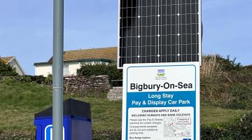 Bigbury car park price board with solar panel and yellow pay here sign on top of a metal pole with a blue waste bin beneath.