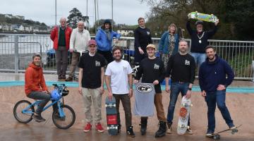 Group of people stand in and around a skatepark bowl, holding skateboards, scooters and one sits on a bmx smiling at the camera