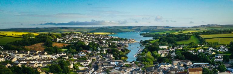 A photo overlooking the town of Kingsbridge from a high elevation. You can see the rooftops of the town, the estuary and the surrounding fields. It is a clear,sunny day with light cloud.